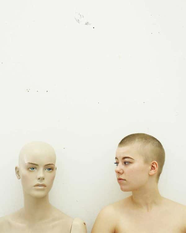 Lara Usherwood - Image of a female model with a shaved head looking at the head and shoulders of a mannequin in front of a white walll