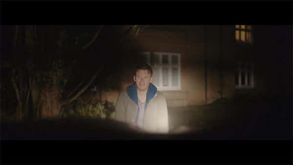 Amnesiac - screenshot from film by Alex Hermon shows a man standing outside of a house lit by the headlights of a car with an upset look on his face and staring towards the camera which is set in the car