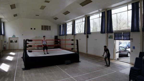 Hitting the Mat - screenshot from film by Alexander Dimmock shows a wide shot of a gym hall during the day with high wide windows and a man walking in through a set of doors in crutches with another man standing in a boxing ring in the center looking at the other man