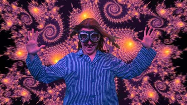 Mr 8 Billion - screenshot from film by James Taylor shows a person wearing blue and white striped baggy pyjamas with open hands raised and aviation goggles on face with long brown hair against bright background of swirling shapes in purple and yellow against a black background