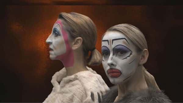 Red Hot - screenshot from film by Josh Dear shows a woman with head turned to camera in white facepaint with overdrawn black eyebrows, eyeshadow and lips with another woman standing behind and looking away with similar white facepaint but with a pink line around head to chin and fur coat