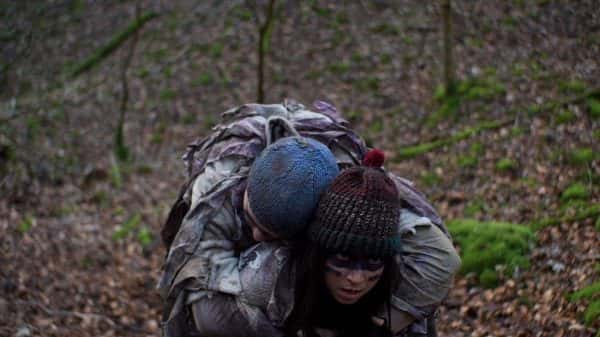 Crawl - Screenshot from student film by Thomas Evans depicts one person wearing a knitted bobble hat and scrappy clothing with painted face around eyes with another person slumped on back wearing similar scrappy clothing in woodland area