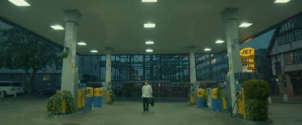 Finding Ferris - screenshot taken from film by Vivienne Warland shoes a man walking through a petrol station pumping with grey sky and fluorescent lights switched on wearing a white shirt and carrying a coat