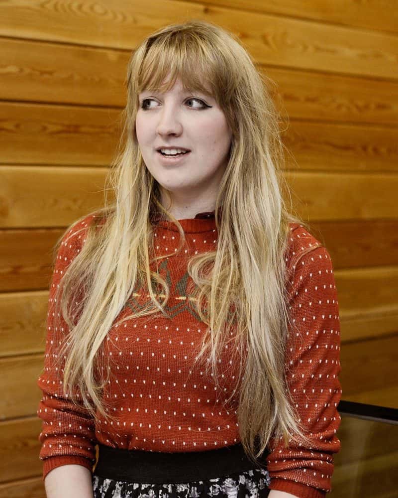 portrait photo of alum Charlotte Simpson smiling and looking off camera with long hair and a rolled sleeve top against wooden wall background