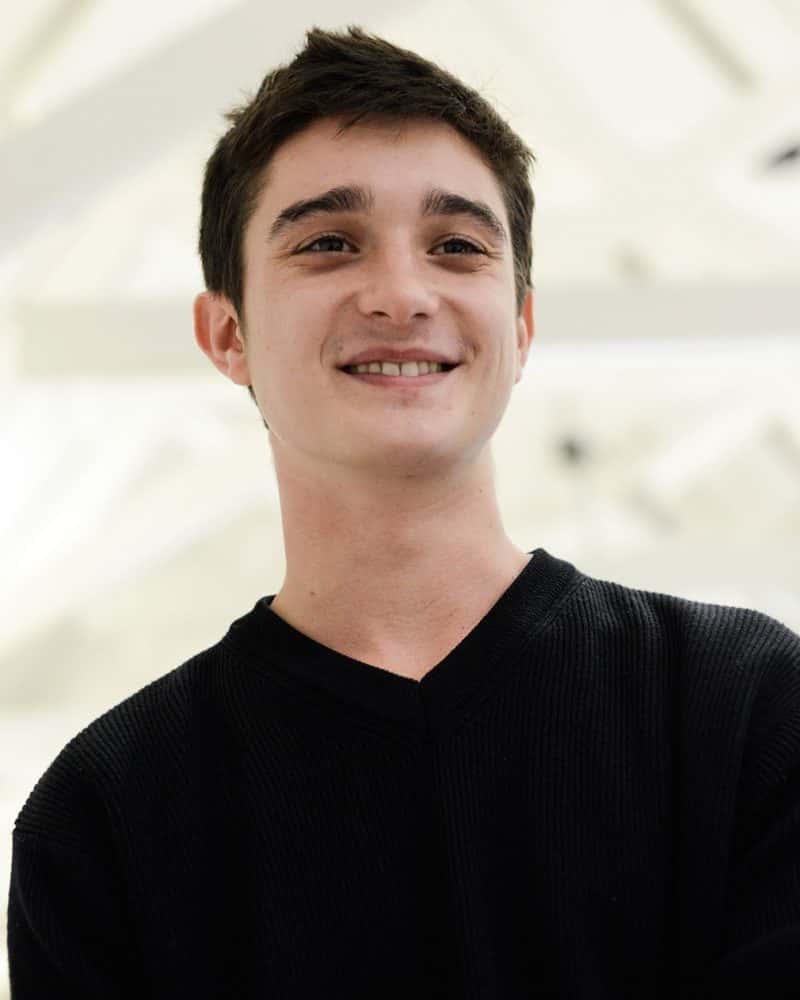 portrait photo alum Stefano Passoni smiling and looking away from camera with short dark hair and a black v neck jumper