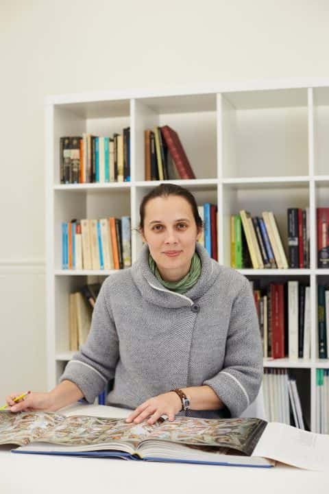 portrait photo of course leader Iuliana Gavril sitting at desk with hands on open book while looking at camera and smiling with tied back brown hair and buttoned grey knit jumper