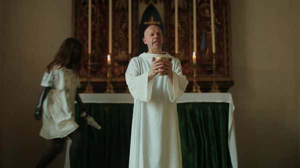 Lake of Fire - screenshot from film by James Rushden shows a man in white robes holding a chalice with both hands and standing in front of a tall alter adorned with candles as another character walks up behind with black shiny skin and a loose white frock dress while holding a small statue