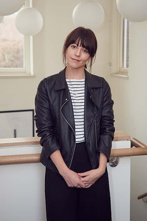 portrait photo of lecturer Kathryn Easthope leaning against wooden railing on white wall and looking at camera while smiling with tied back hair and open black leather jacket