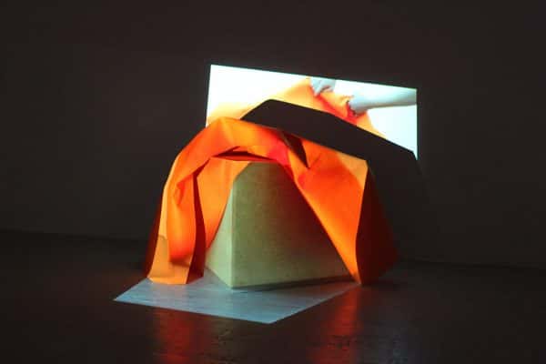  - photo of exhibition piece shows lit cube with orange fabric draped over the top and a projection showing hands manipulating the same cloth being played over it