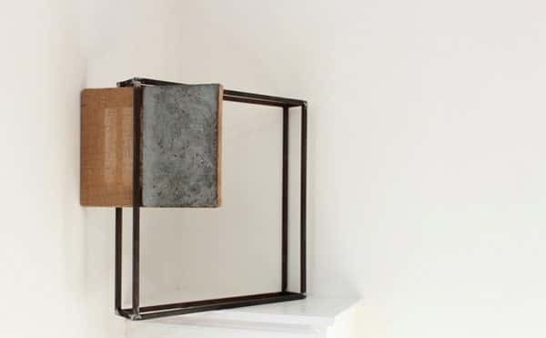  - photo of exhibit piece shows white wall with white table holding up a black metal cube frame with a split fabric and grey wood board suspending at an angle in one corner