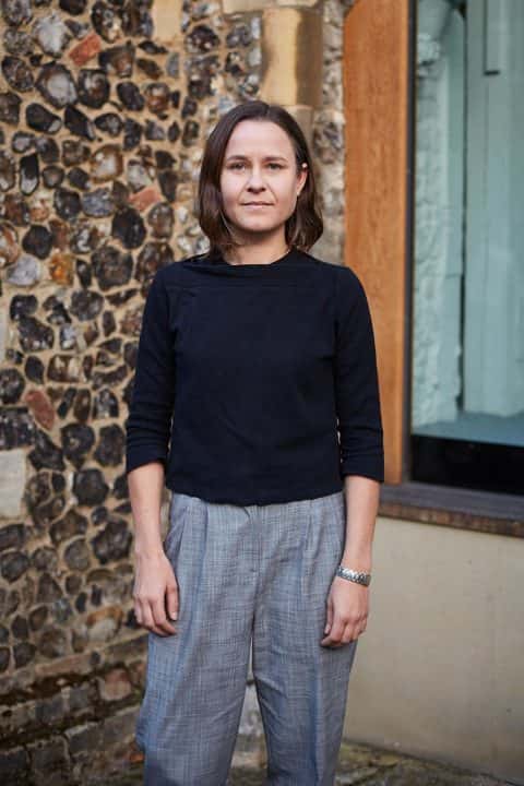 Portrait photo of lecturer Gretchen Geraets standing with arms by side and smiling at camera with medium brown hair and a rolled sleeved dark blue jumper
