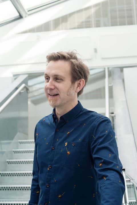 portrait photo of senior lecturer Glen Robinson standing and looking away from camera with short light brown hair and a long sleeved blue shirt yellow splash patterns