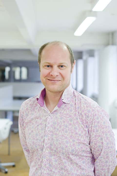 portrait photo of course leader Glyn Brewerton standing with arms behind back and smiling at camera with short blonde hair and a pink floral long sleeved shirt