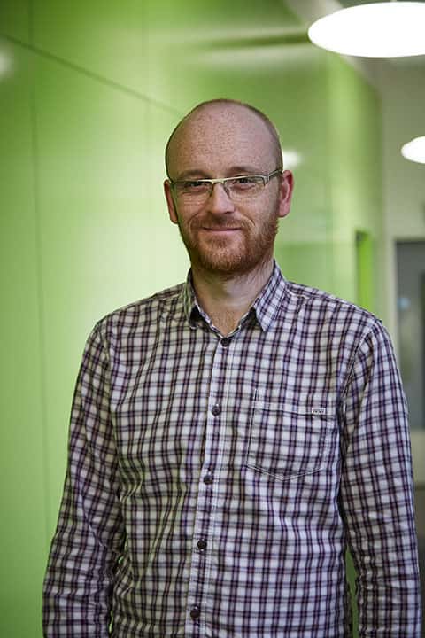 portrait photo of lecturer Jon Dunleavy standing with arms by side and smiling at camera with short red hair and a plaid white and red shirt