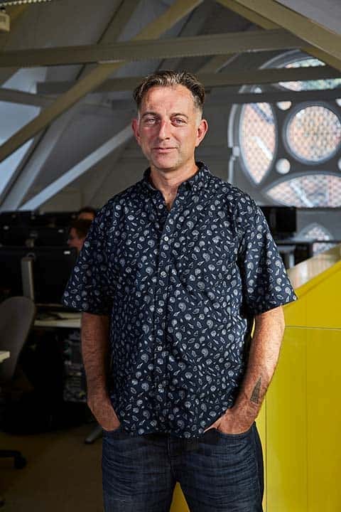 photo of Lecturer Jamie Gledhill standing with hands in pockets and smiling at camera with short light brown hair and a loose patterned blue shirt