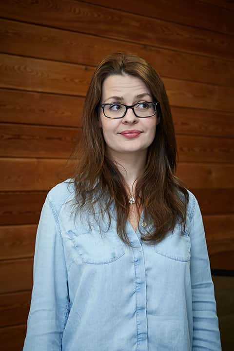 portrait photo of lecturer Katy Hammond standing and looking away from camera with long brown hair and black glasses and light blue denim shirt
