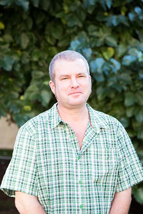 portrait photo of senior lecturer Les Bicknell standing with hands crossed and smiling at camera with short grey hair and loose green and white plaid shirt