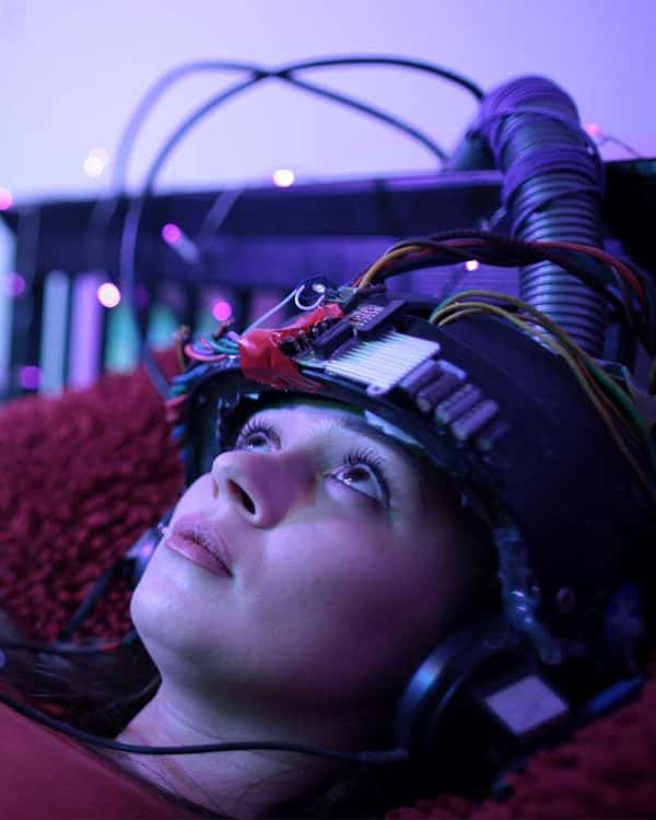 Luke Green - Still image of a girl laying on a bed, she has a helmet on which is connected to many wires and she is staring up at the ceiling