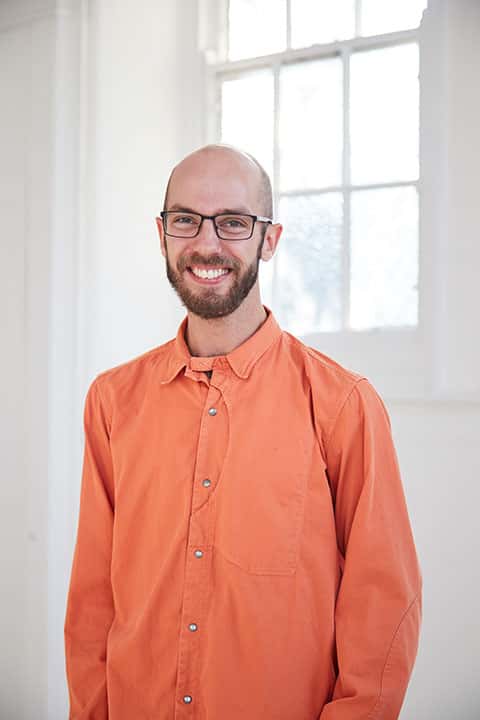 portrait photo of lecturer Matthew Benington standing with arms at side and smiling at camera with short brown hair with black square framed glasses and wearing an orange long sleeved loose shirt