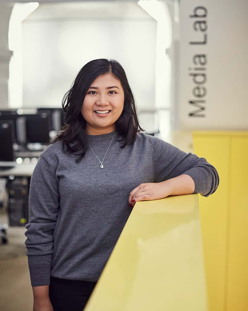 photo of alum Leana Felipe standing and leaning against a yellow railing and smiling at camera with long black hair and a long sleeved grey top in a room with many computer benches in the background