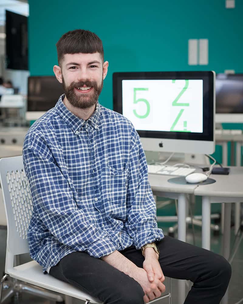 photo of alum Matthew Longley sitting on a white chair and smiling at camera with hands crossed in lap and short brown hair wearing a loose blue and white checked buttoned shirt with several computers on benches in background