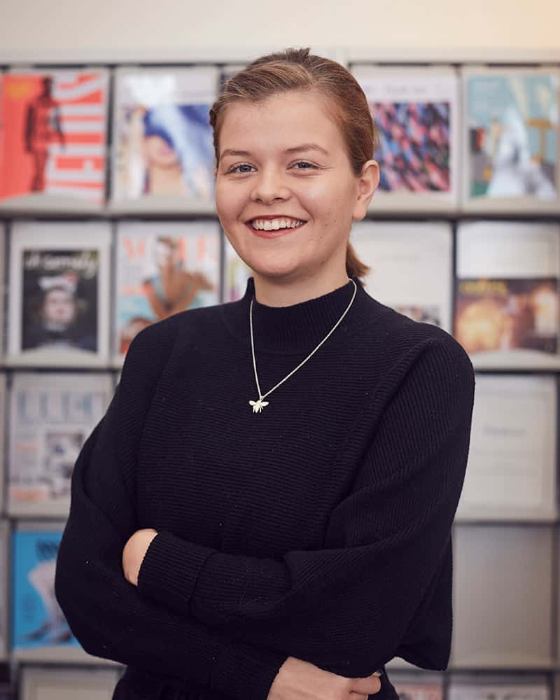 portrait photo of alum Faye Anderson standing and smiling at camera with tied back hair and a long sleeved black jumper in a room with a rack of design work in the background