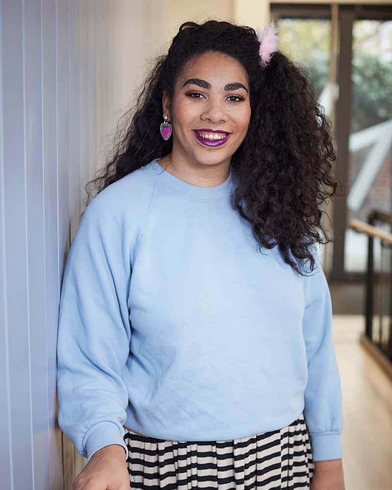 photo of alum Florence Watson standing next to wall with arm on railing and smiling at camera with long curly black hair and a loose light blue jumper in a tall room with glass doors in background