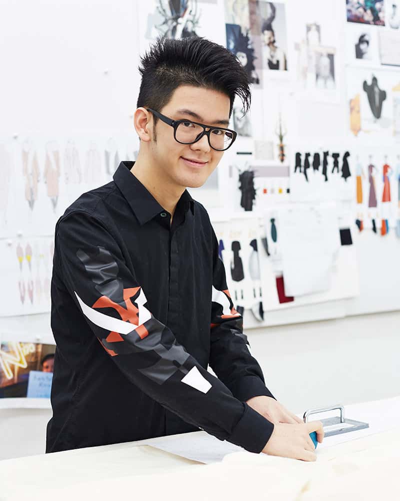 photo of alum Jerry Huang standing at design desk and smiling at camera with hands on fabric material and spiked black hair and thick black glasses and black long sleeved buttoned shirt in a room with a large board on the wall covered with design work