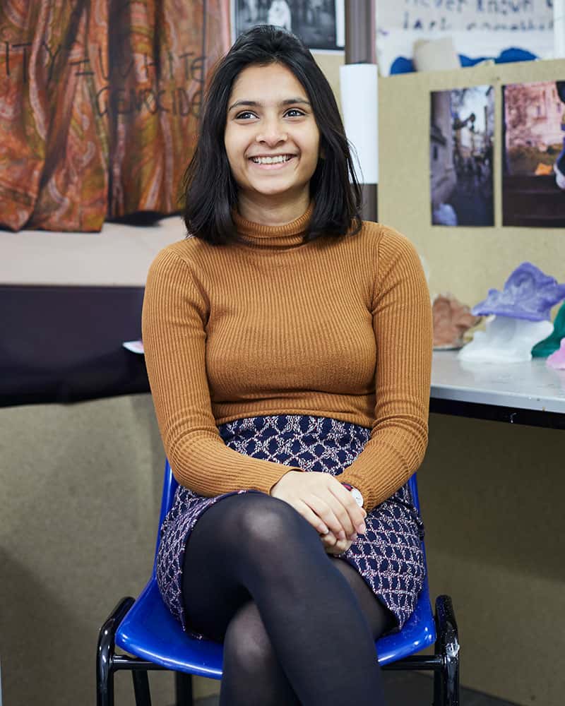 portrait photo of alum Alveera Rahman sitting in chair and smiling away from camera with black medium hair and brown long sleeved jumper