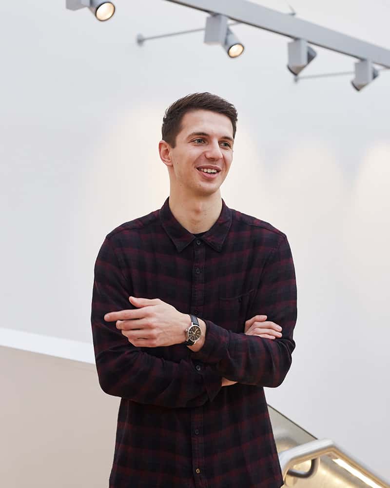 photo of alum Craig Riseborough standing and looking away from camera with short dark brown hair and wearing a checkered red and black shirt with a white wall background
