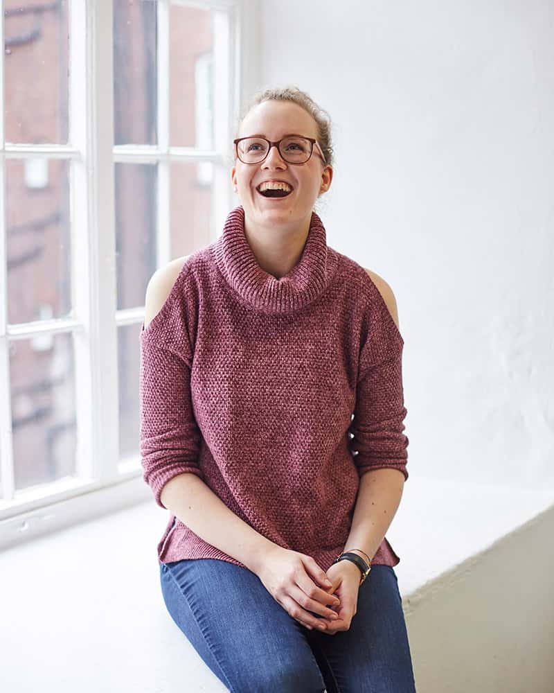 photo of alum leoni lessmann sitting on a ledge next to a window and smiling whilst looking up and with tied back hair and glasses with a rolled neck knitted jumper and crossed hands