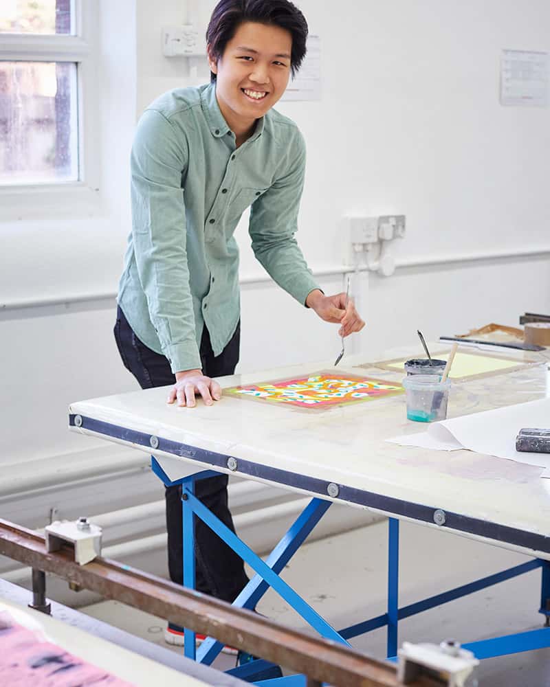 photo of alum Brandon Tan leaning over a desk and holding a spoon and smiling at camera with swept black hair and a denim buttoned shirt in a design studio with white walls and a window