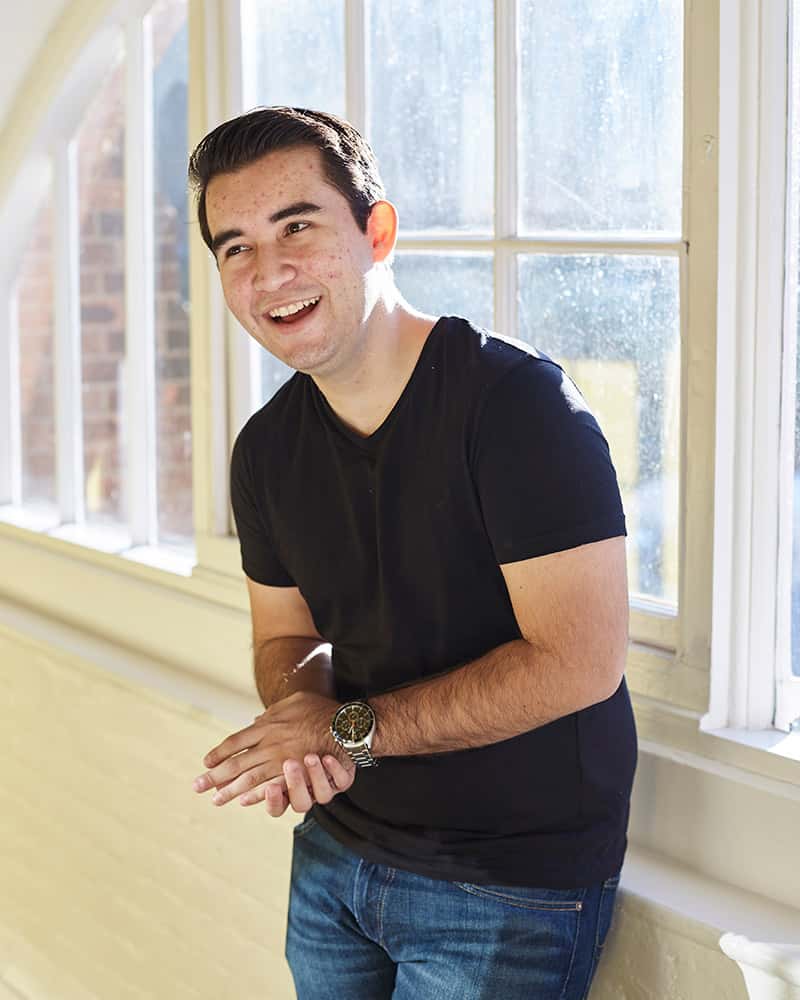 photo of alum Jose Fernandez looking up and smiling with combed black hair and wearing a black t-shirt with a wide bright window in the background