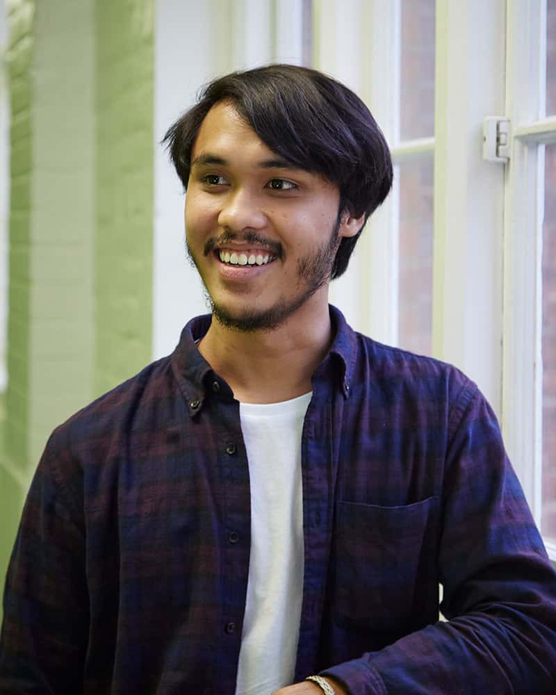 photo of alum Fahim Fadzlishah smiling and looking away from camera with arm leant against wall and short swept black hair and an open checkered red and blue shirt with window in background