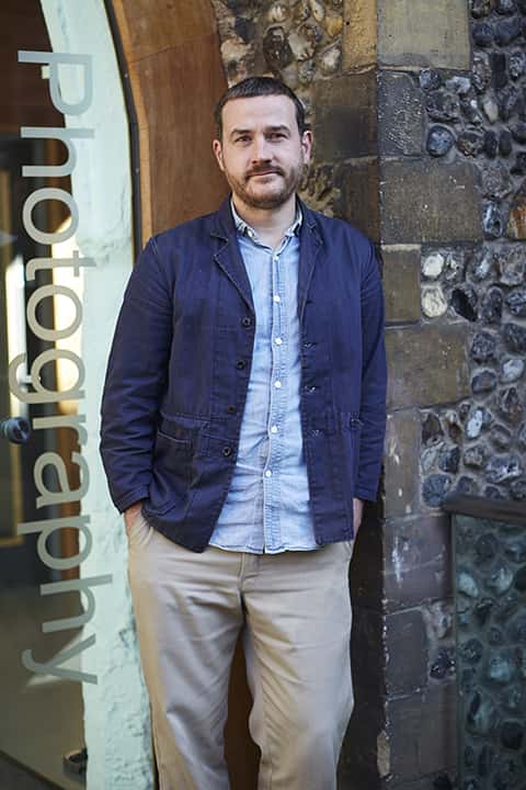 portrait photo of lecturer Ross Trevail standing and leaning against a stone wall with short black hair and a blue open denim jacket