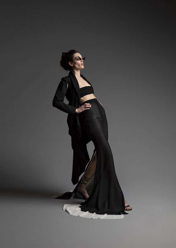 Angela Celestino Duarte - Model mid-stride with hands on hips leans back dramatically, wearing sunglasses with slanted frames, an open long black jacket, and a long black split-side skirt with white rim