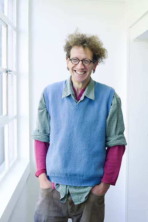 portrait photo of lecturer Nigel Bousfield standing with hands in pockets and smiling at camera with curly blonde hair and round brown glasses with a blue sweatervest and plaid green and white rolled sleeve shirt and a long sleeve pink undershirt