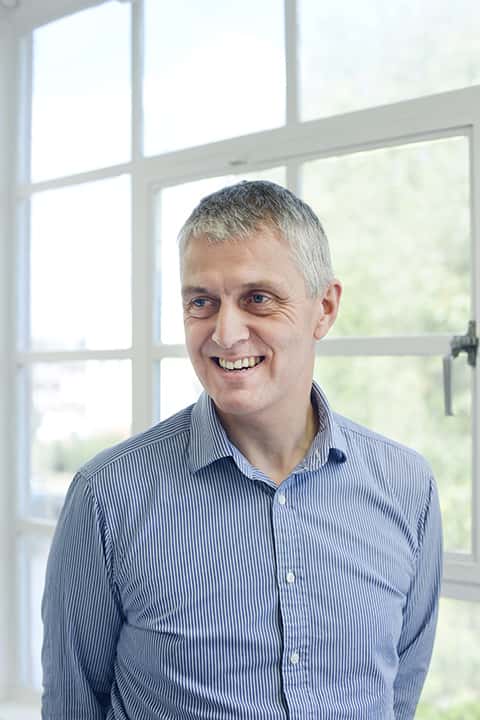 portrait photo of lecturer Nigel Coton standing and smiling away from camera with short white hair and a vertically striped white and blue long sleeved shirt