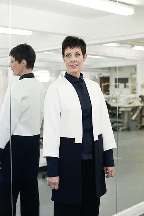 portrait photo of associate professor and course leader Sue Chowles standing next to tall wall of mirrors and smiling whilst looking away from camera with short black hair and a white and blue coat jacket