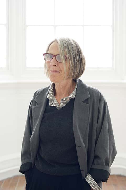 portrait photo of Research Supervisor Victoria Mitchell standing with hands in pockets and smiling at camera with medium blonde hair and red glasses and a grey loose open shirt jacket
