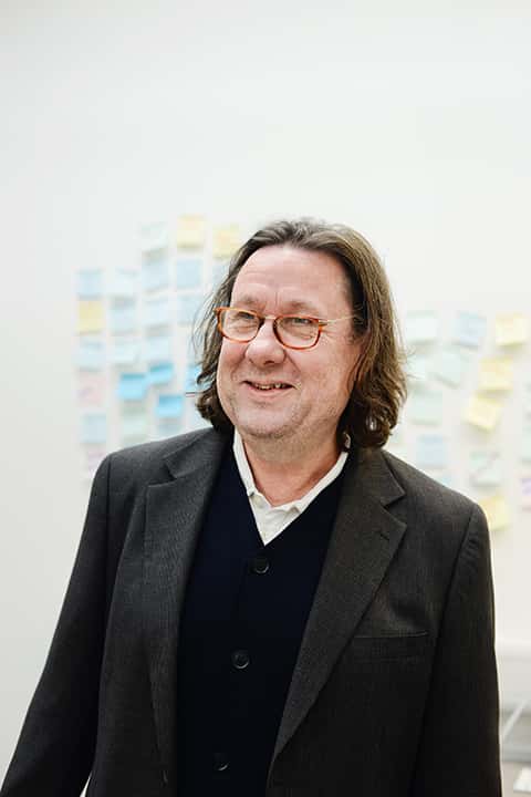 portrait photo of Dr Rob Hillier standing and smiling while looking away from camera with medium brown hair and brown glasses with a dark grey suit jacket