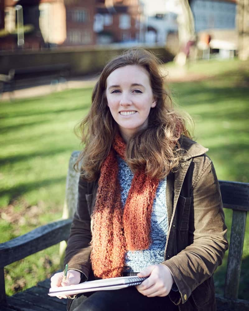 photo of alum Kaelin O Hare sitting on wooden bench in grassy park and holding sketchbook while smiling at camera with long brown hair and a knitted scarf and a medium length green coat
