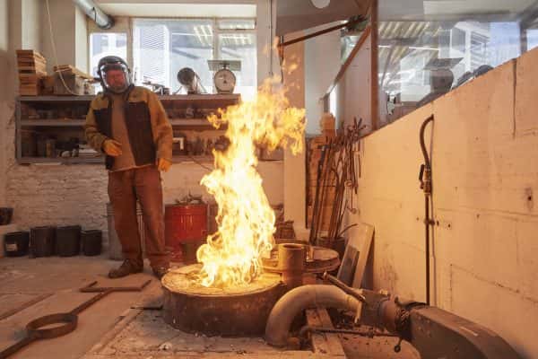  - A technician doing a demonstration of 'the pour' in the foundry in the 3d workshop watches a large (controlled) flame while pouring molten metal to make a sculpture