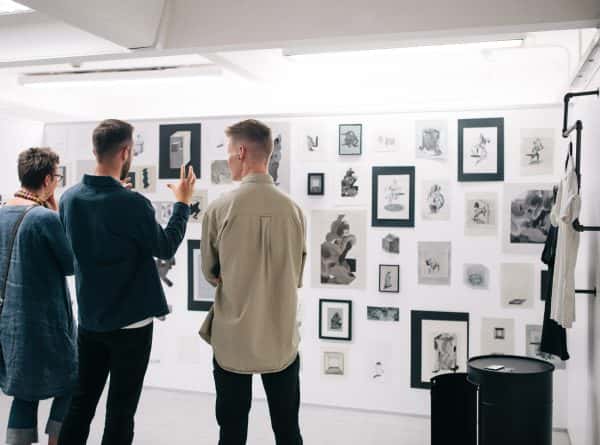  - Three people looking at a wall full of framed drawings in the 2017 BA Degree Show