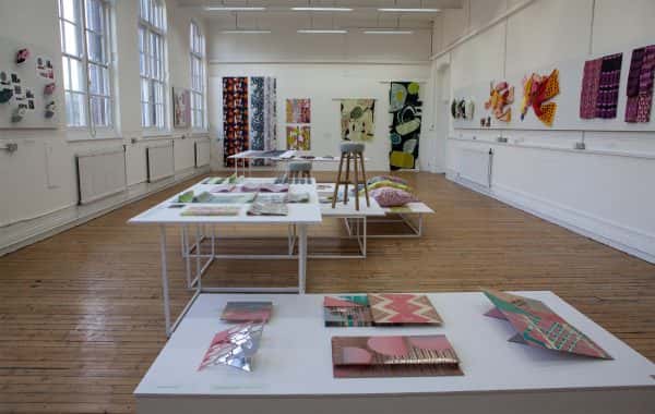  - Large room with wooden floor displaying textile design work on tables and walls in 2017 BA Degree Show