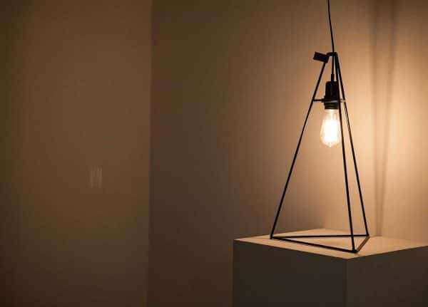  - Image of a metal framed lamp in a dark studio space, the lamp is lit and shines a yellow light into the space