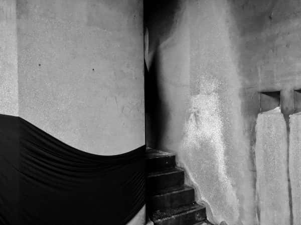 Emily Cannell - Monochrome image of a set of steps emerging from close concrete walls with black fabric draped along the wall away from the stairs