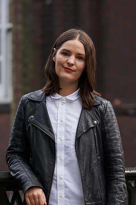 BA Fashion Marketing and Business Lecturer Holly Farrar stands for a profile photo in a leather jacket and white shirt, smiling at the camera