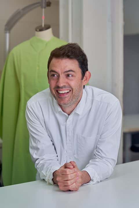 Eugene Reeder, BA Fashion Senior Lecturer at Norwich University of the Arts smiling and leaning on a bench in front of a fashion mannequin with a green garment on