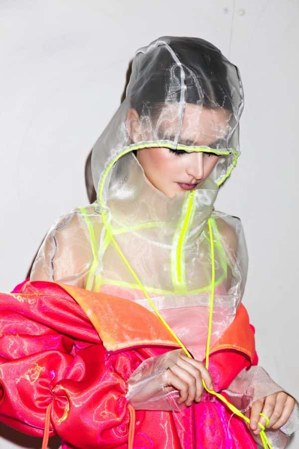 Anya Sims - White female model wears a layered neon pink, yellow and orange jacket, partially unzipped. Underneath is a transparent grey and neon yellow hooded top. Designed by BA Fashion student Anya Sims